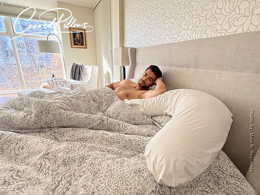 A shirtless model lying in bed, positioned atop a curved pillow. The image is featured in a blog post titled ‘There’s Only One Real Benefit of Sleeping Naked.